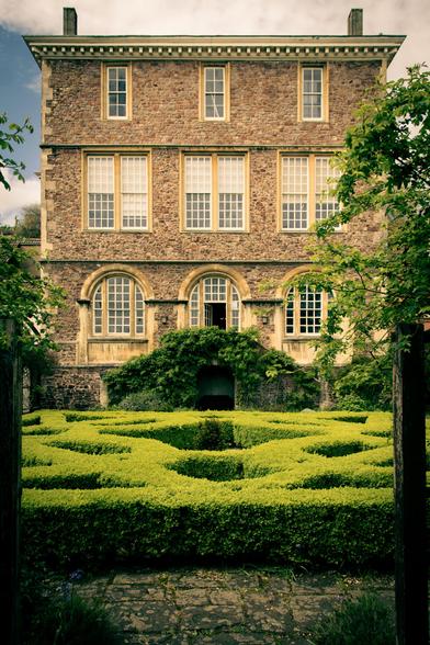 View of a historic house from across the garden with boxed hedges: ivy-covered stairs lead to the door on the ground floor.