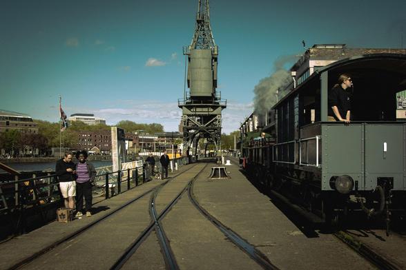 A quayside view in the sunshine: train tracks, a historic crane, and the final waggon on a stream train, as well as people milling about.