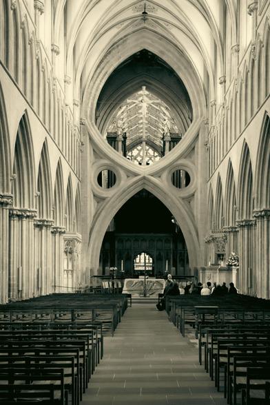 Black and white view along the nave of this gothic cathedral towards the altar. The lines of the architecture move the eye upward as well as straight ahead, where the famous scissor arches appear.  Some people on a guided tour are sitting inthe front chairs on the right.