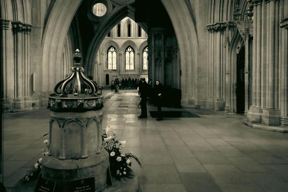 Black and white photo of the interior of the gothic cathedral in Wells. From the baptismal font in the foreground leads you to some tourists milling around and then through the famous scissor arches to the stained-glass windows at the north end.