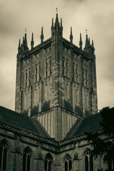A monochrome image of Wells Cathedral. Above the corner where the roofs meet, the squat, square spire rises against a cloudy sky.