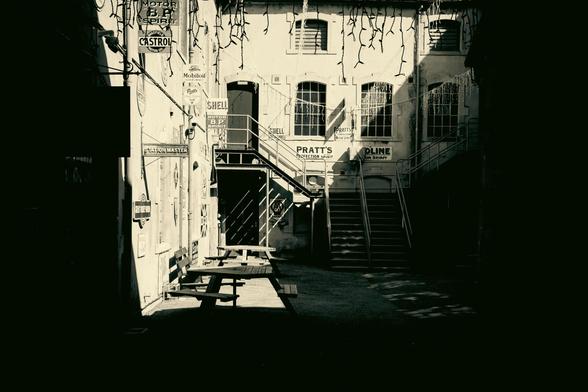 A black and white image of an old industrial building in bright sunlight and a dark shadow around the edges. Visible are some stairs leading to a door on the first floor and several windows. The walls are decorated with old advertising signs.