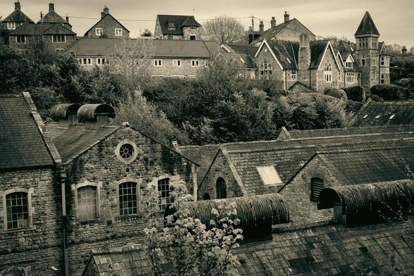 Black and white image of a view over the rooftops of an old industrial building looking towards a series of houses (one Victorian with a turret) higher up on the hill.