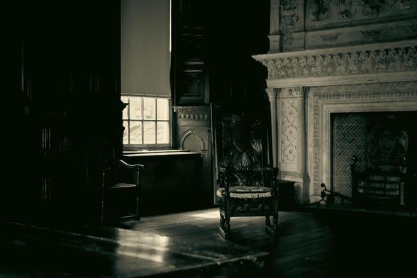 Black an white image of a grand room inside a historic house. The blind is half drawn in front of the window. In the shades are two antique chairs and a large fire place and decorative chimney breast on the right.