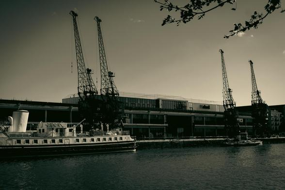 A black and white view of the Bristol harbour. Across the river, the Balmoral is moored at the quay; behind it are two pairs of historic cranes and the building of M Shed, the famous museum.