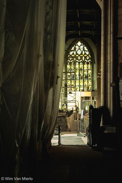 Interior of the church while constructions work is going on.  We are looking towards the light of a stained-glass window while the foreground is in darkness; drapes on the left are protecting some of the church interior from dust.
