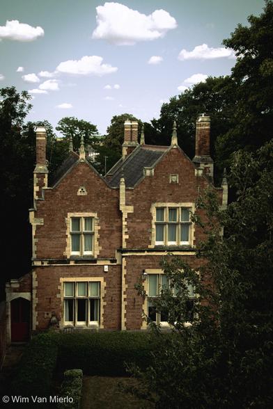 An old red-brick building with four large windows and several chimneys surrounded by trees underneath a blue sky with scattered white clouds.