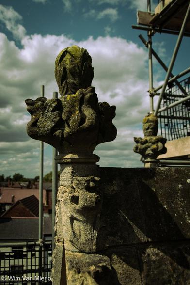 A well-weathered stone carving on top of a church roof. In the background is scaffolding and a blue sky with big, white clouds.