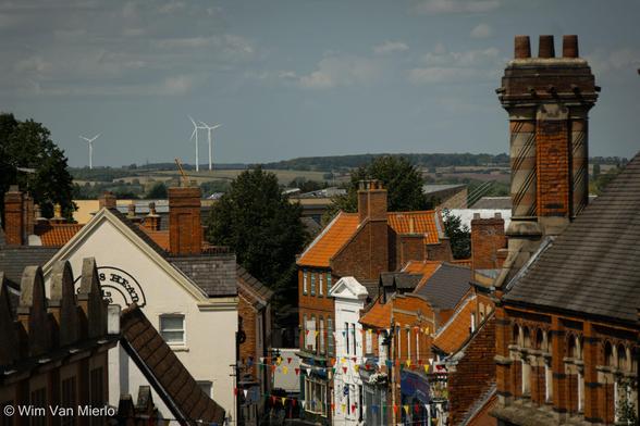 View across the rooftops of the town, showing red and white houses in different styles; a large building with a interesting double chimneystack; on the horizon are three windmills generating clean energy.