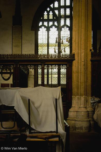 A church interior: some of the furniture is covered in a white sheet to protect it from dust; a stone pillar, wooden screen, and arched window.