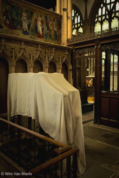 A church interior: some of the furniture is covered in a white sheet to protect it from dust. Above this is some arched stonework and a religious painting; to the right a wooden screen with doorway leading to arched windows.