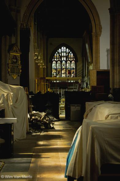 View of the interior of a church towards a stained-glass window; various items of furniture are covered in white sheets to protect them from dust; the sunlight ins pouring in from the left.