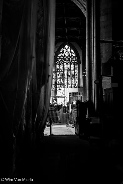 Black and white interior of a church with heavy shadows during construction. Looking in between white sheets draped from ceiling to floor on the left and a stone pillar on the right towards a stained-glass window.