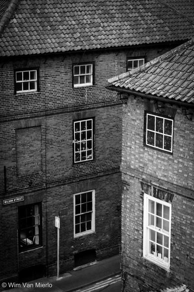 Black and white image of a streetcorner with two brick houses of an earlier period with white-framed windows and tiled roofs. A traffic sign is below the streetsign, which reads 'Wilson Street'.