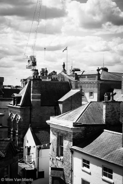 Black and white image of a view across the town's rooftops. Behind a mixture of hose in different styles sticks out the framework of crane on a construction site.