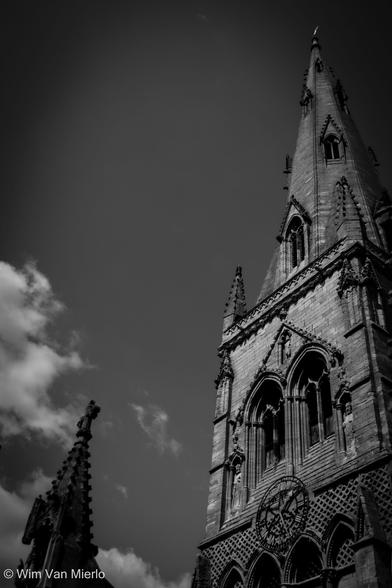 Black and white image of a church spire of a mediaeval church under a blue sky with scattered clouds; a smaller turret is to the left of the clock face and arched windows.