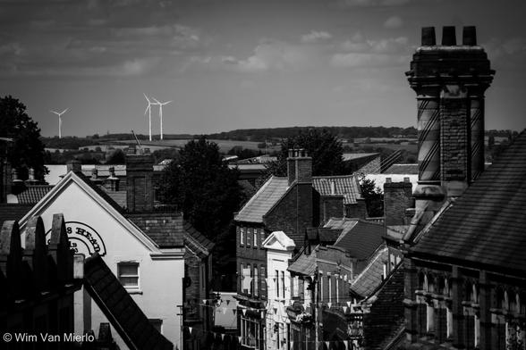 A black and white image across the rooftops of an English town on a bright day.  Houses in various styles: on the right is an impressive triple chimney-stack. Three windmills generating green energy are visible near the horizon, where part of the surrounding country-side is visible.