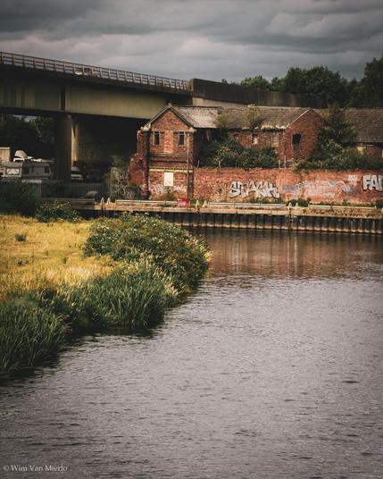 A bend in the river passed some grassland on the left an an abandoned red-brick building and wall further back.  A concrete viaduct carries an A road across. 