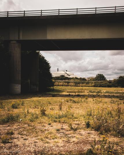 An empty over which a viaduct carrying an A road crosses. In the distances, the white top of a factory is visible.