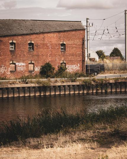 At the quay on the other side of the river stands an abandoned, red-brick warehouse with graffiti on the walls. To the right is a gantry of the railway running alongside.