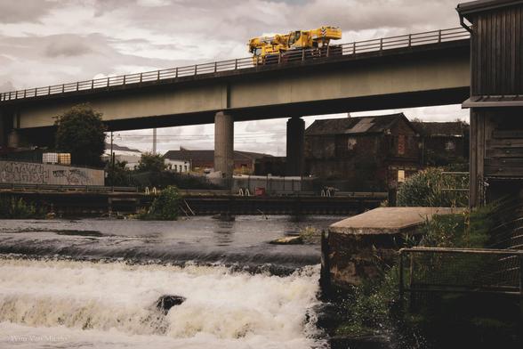 An A-road crossing the river Trent at a weir; a yellow vehicle is driving past on the concrete viaduct under a cloudy sky.
