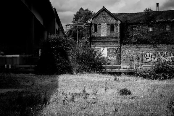 A black and white image with high texture of an old industrial building covered in graffiti alongside a a road viaduct in the shadows.