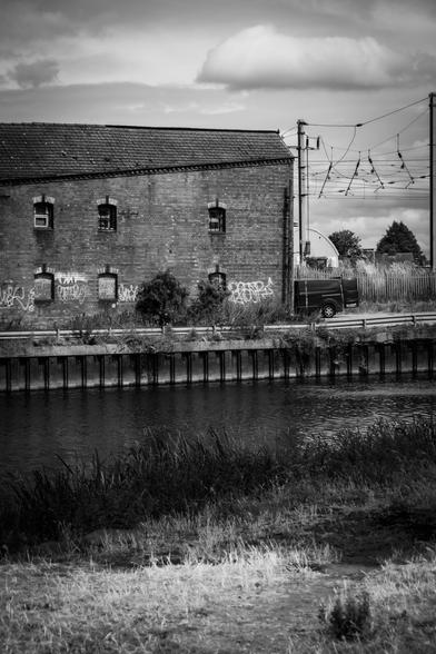 A black and white image of an old warehouse along the river quay; the gantry of a railway line passing behind is seen on the right.