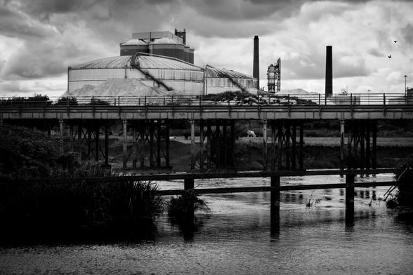 An industrial scene with the silos, domes and chimneys of a factory appearing above the railway bridge crossing the river Trent.