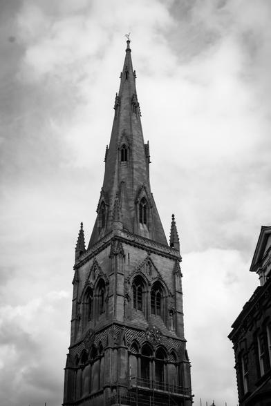 Black and white, somewhat moody image of the top half of a gothic church spire against a partially cloudy sky.