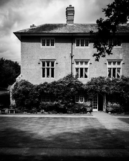 Black and white image of a stately manor wit ivy on the walls; only the left side of the house is visible across the lawn; strong contrast between sunlight and shadows.