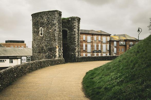 Photograph of old tower, part of the city wall; mixture of building sin the background; a grass mound on the right; grey sky.
