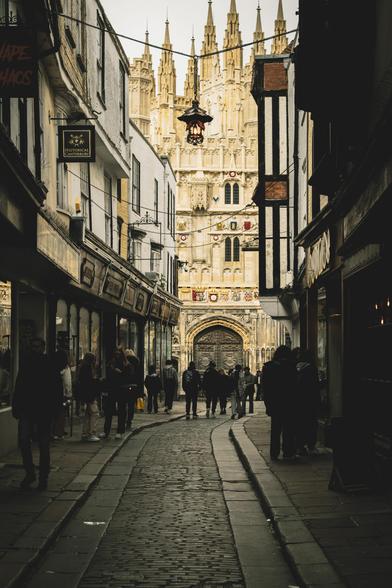 View down a narrow, shadowy street with people walking towards the bright and impressive gothic frontage of Canterbury Cathedral.