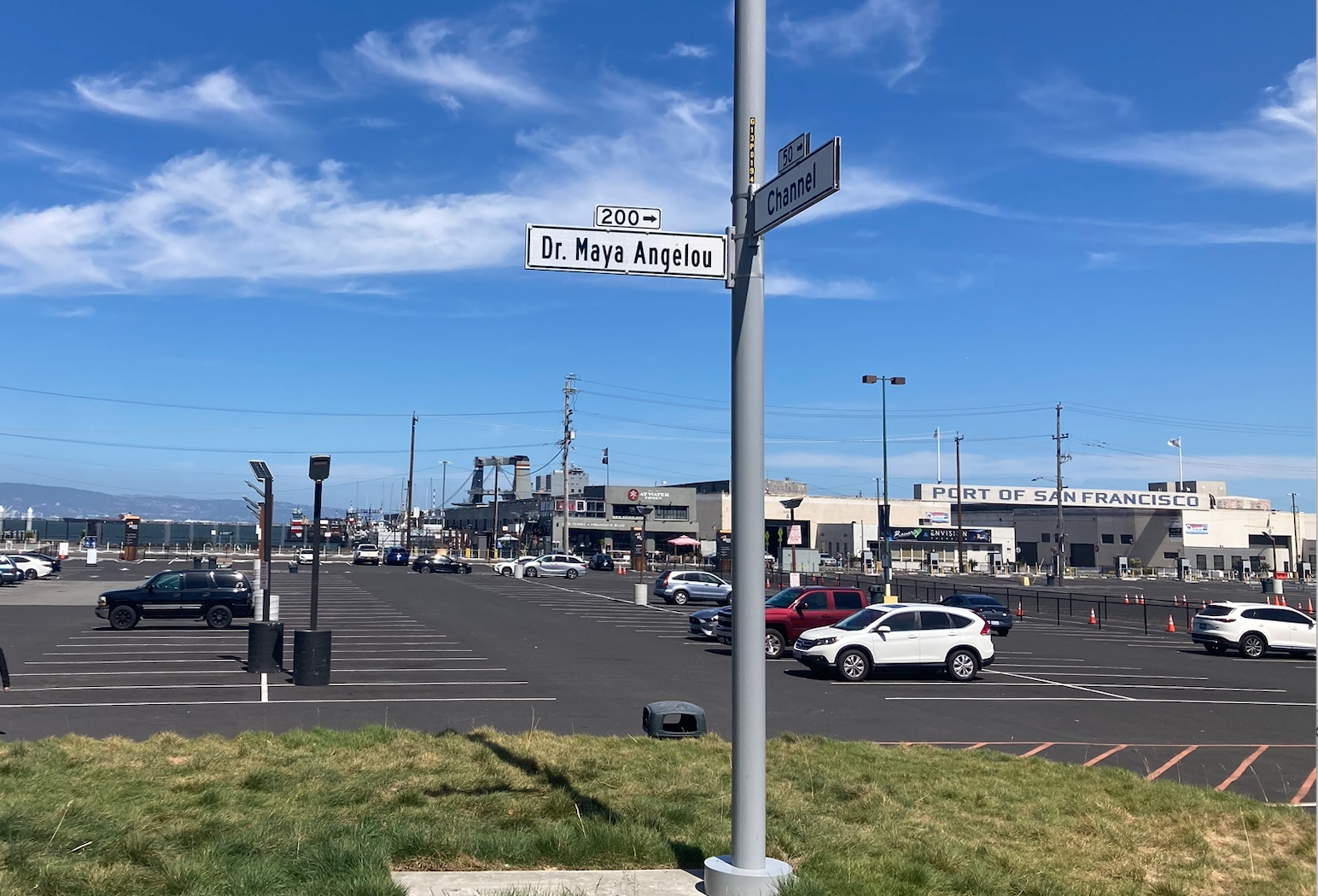 Street sign, Dr. Maya Angelou. In background is a beautiful blue sky with wispy cloud and a parking lot with some cars.