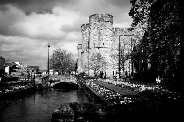 A black and white image of a river leading to a stone bridge and the medieval towers of the old city gate.
