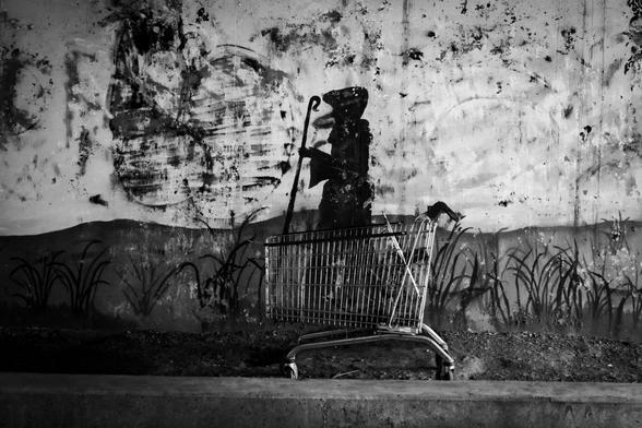 Black and white photo of an an abandoned shopping cart in from of graffiti depicting a pilgrim.