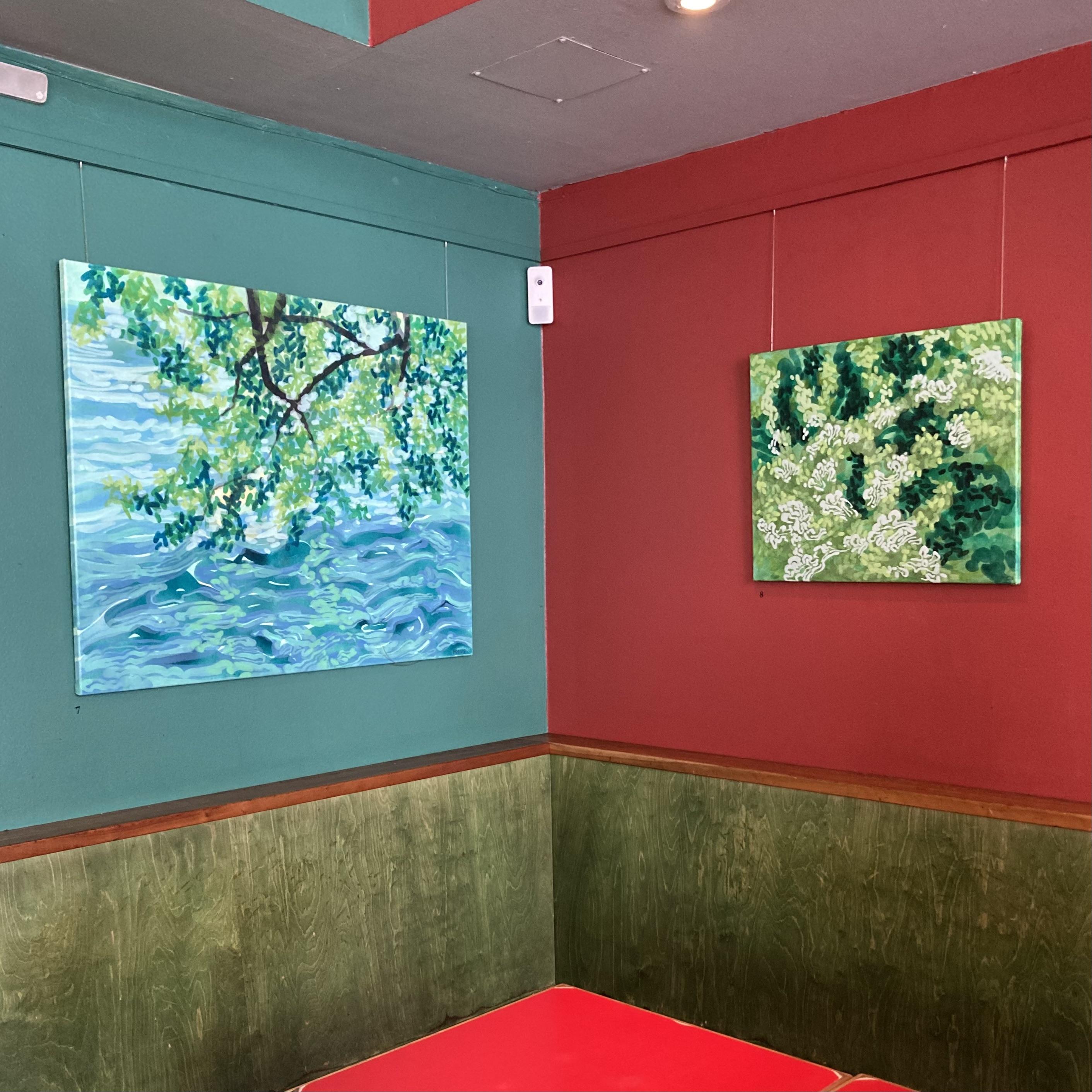 A interior shot inside the café of a corner with a painting on the left and right walls. The painting on the left depicts an abstracted scene of floodwater high enough to reach the green boughs of a a tree. The painting on the right is an abstract composition in shades of green representing turbulent water.