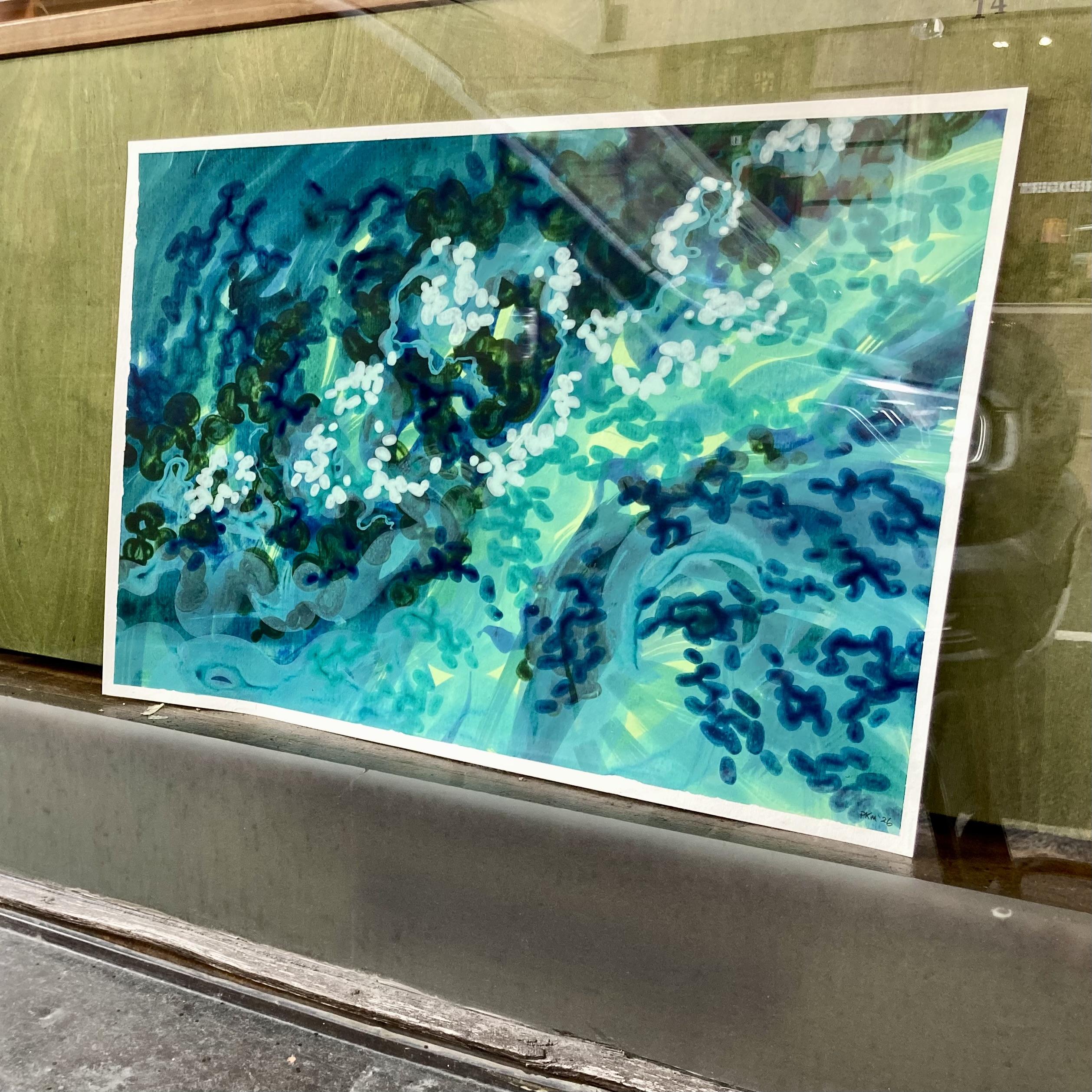A view from outside a café window looking in at an abstract painting on paper that is propped up behind the glass. It has a swirling composition in shades of teal, green, and blue that depicts water currents.