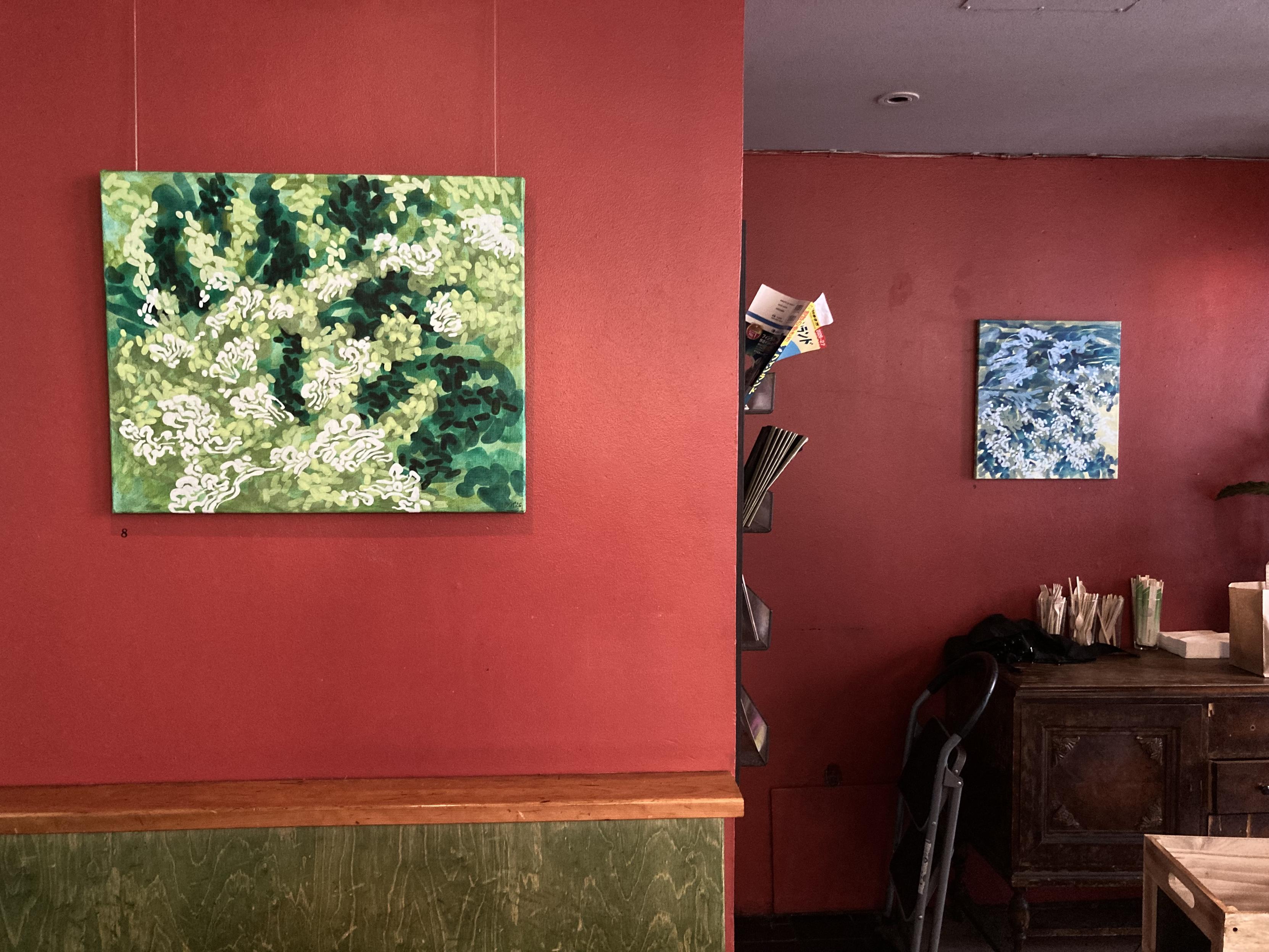 A interior shot inside the café of two walls with a painting on the left near and right far walls. The walls are painted deep red. The painting on the left is an abstract composition in shades of green representing turbulent water. The painting on the right is a mostly blue and off white abstract composition of water flowing in river rapids.