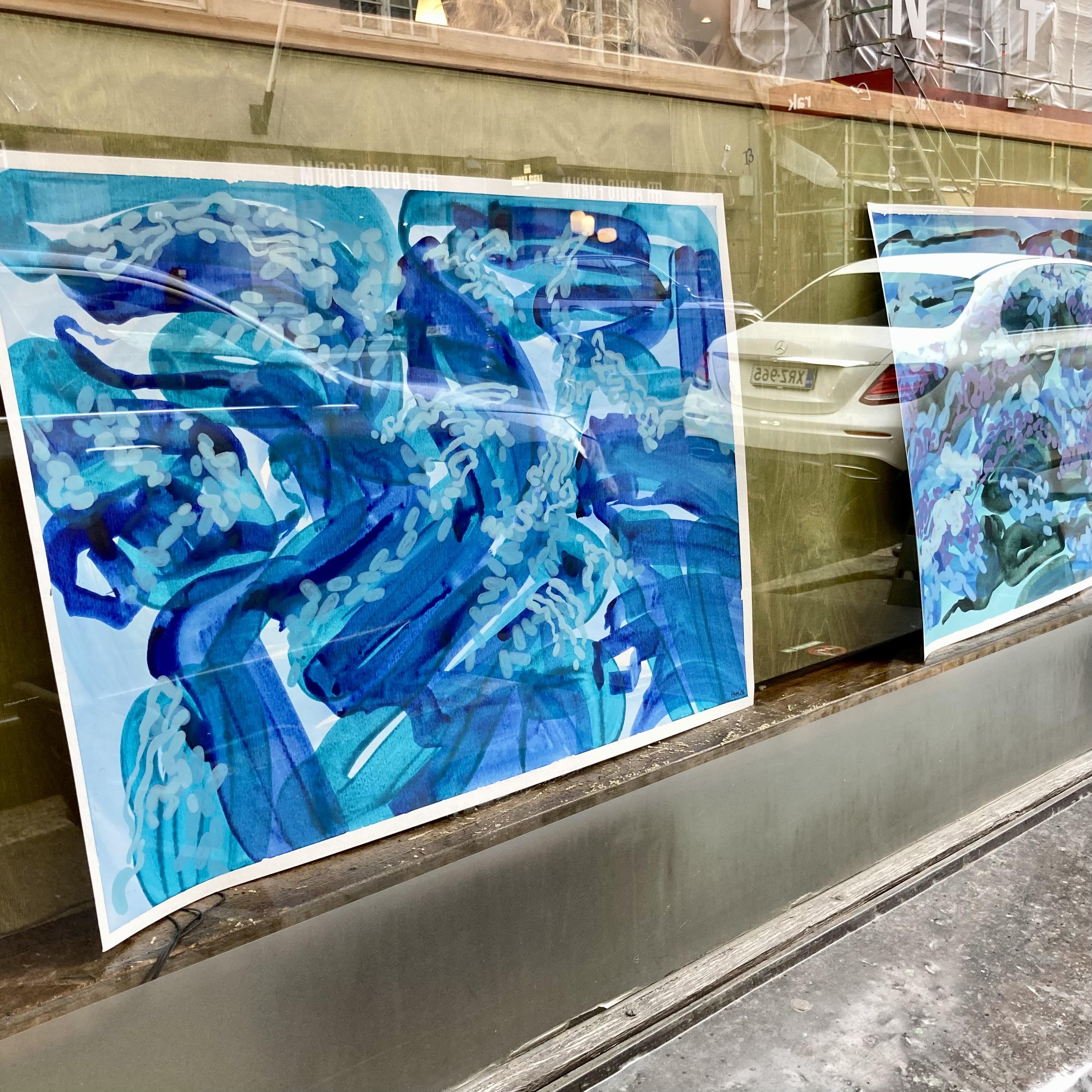 A view from outside a café window looking in at two abstract paintings on paper that are propped up behind the glass. The paintings have compositions in shades of teal and blue that depicts moving water.