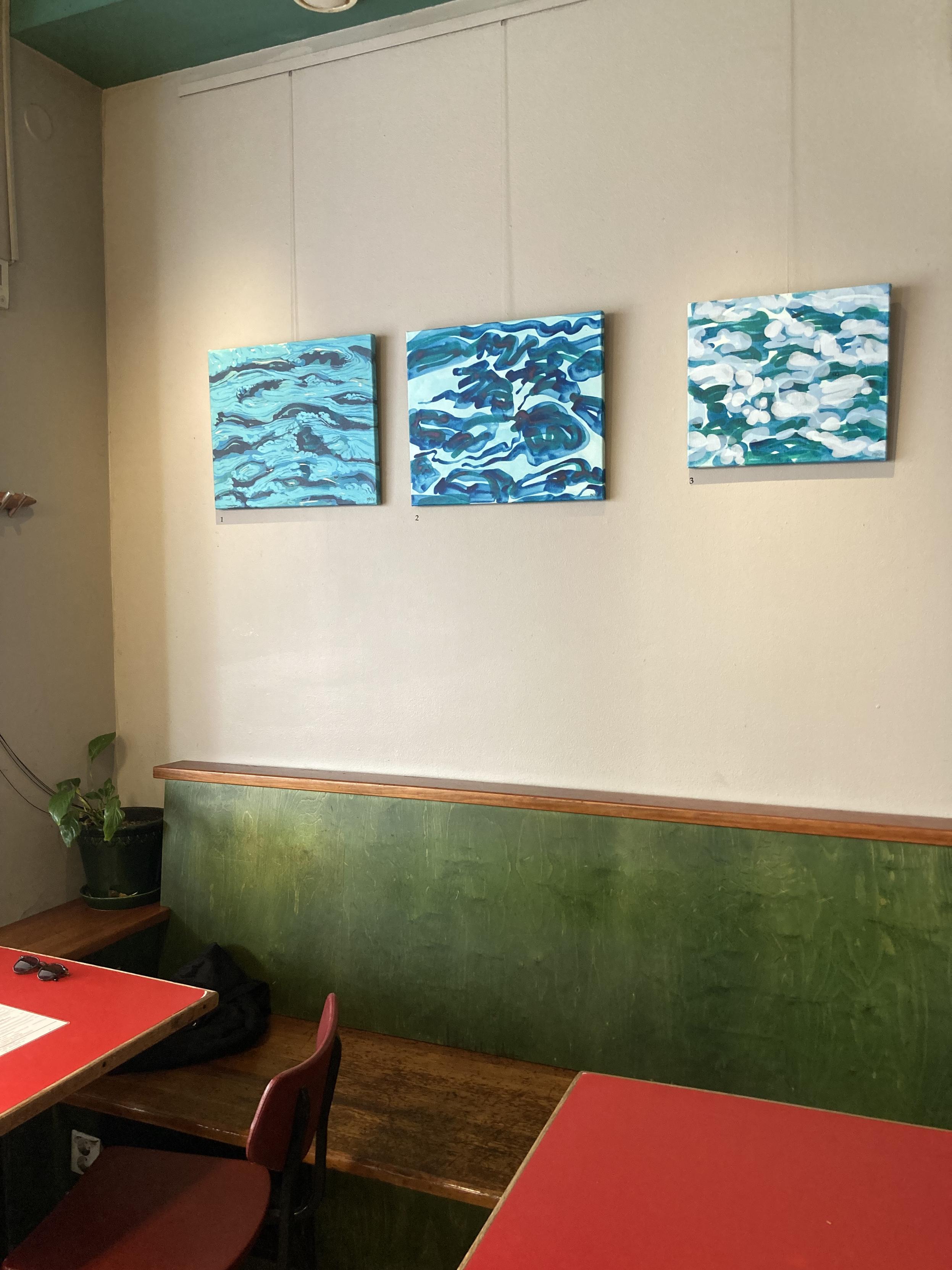 A interior shot inside the café with a cream coloured wall behind sap green benches and red table tops. There are three small abstracted paintings hanging close together on the wall. The paintings have a colour palette of blue and teal and represent waves in moving water.