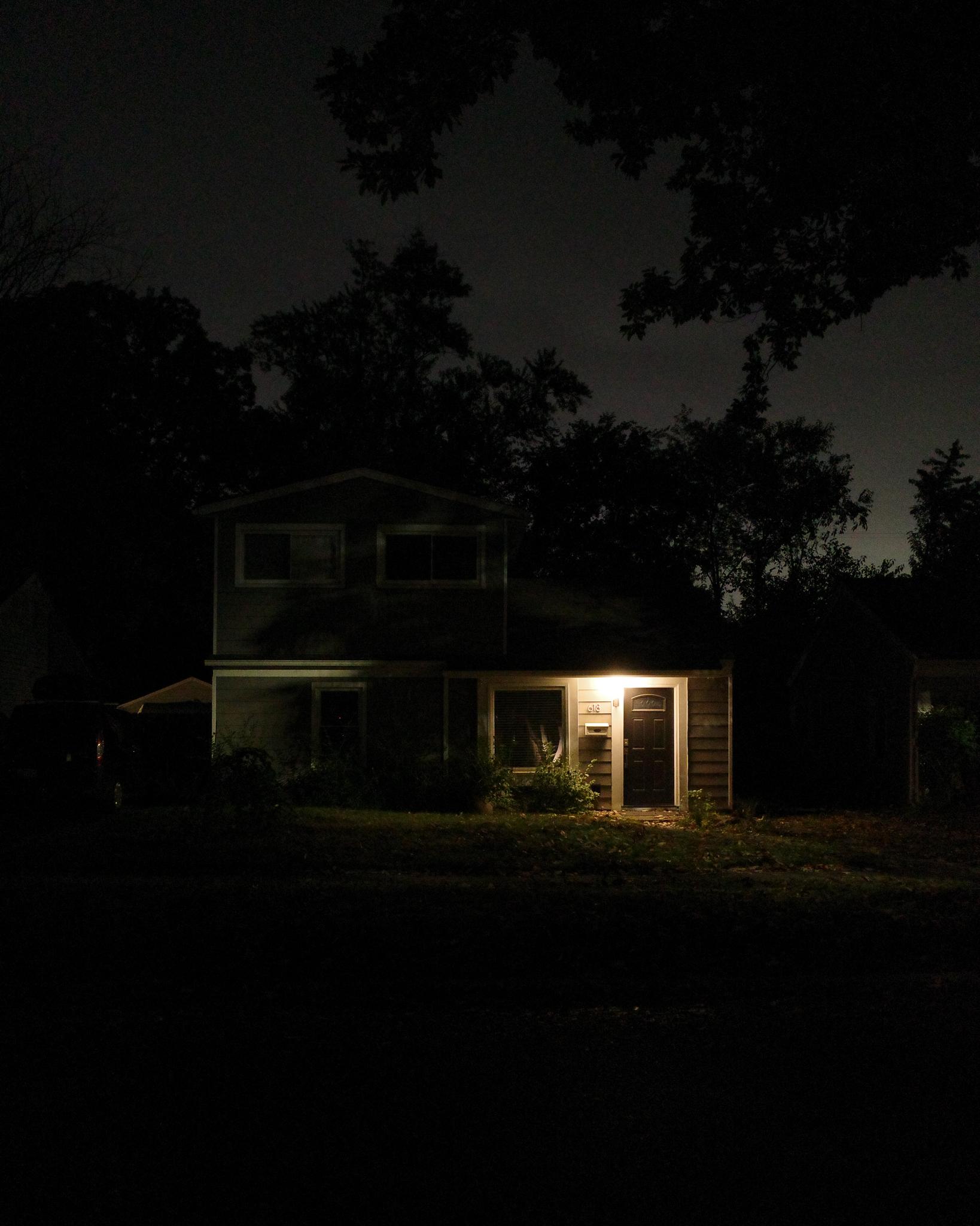Color photograph of a darkened house at night, tree branches around it silhouetted against a glowing night sky