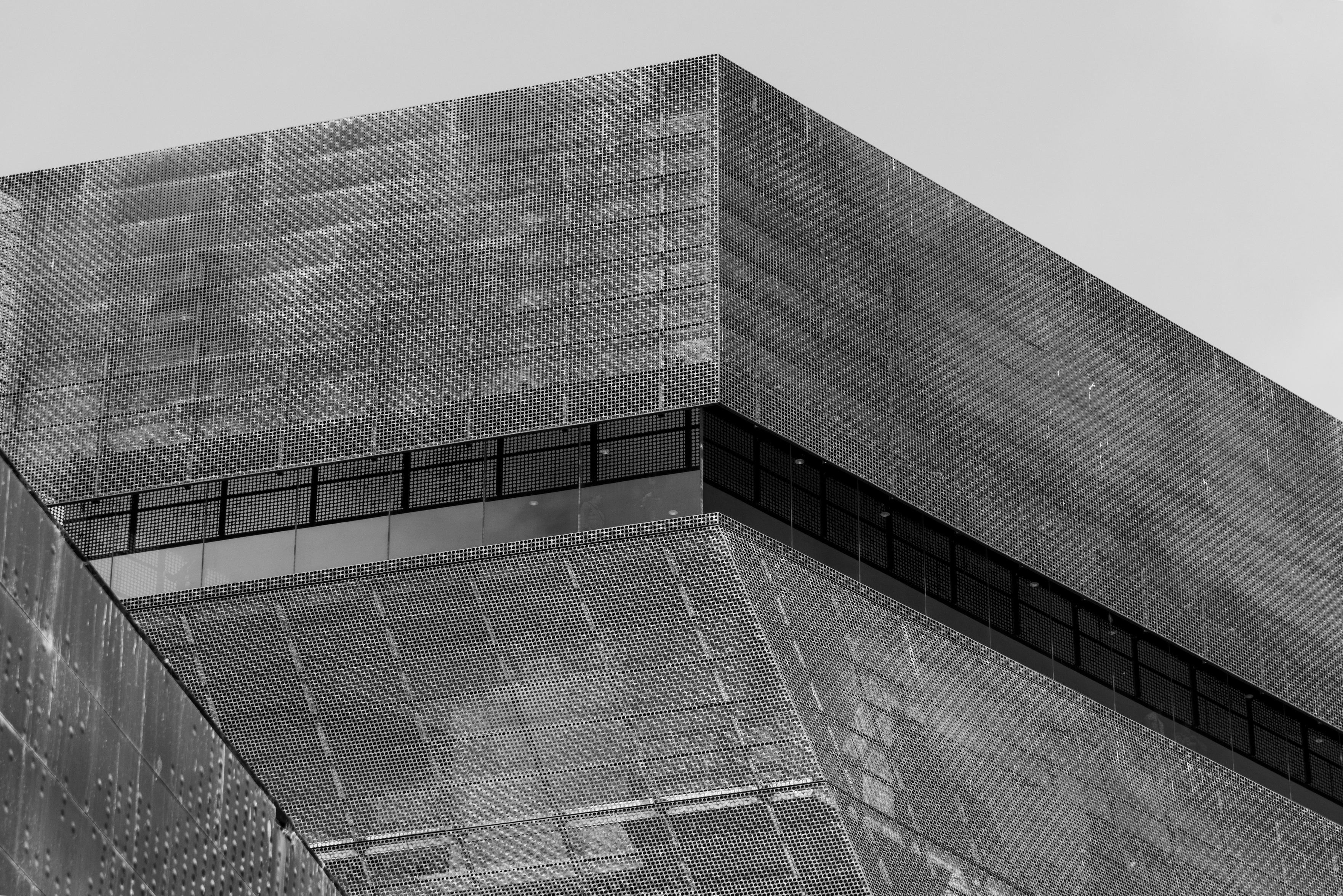 Black and white photo of a tall, cantilevered structure - clad in perforated copper panels - which features a single wraparound window