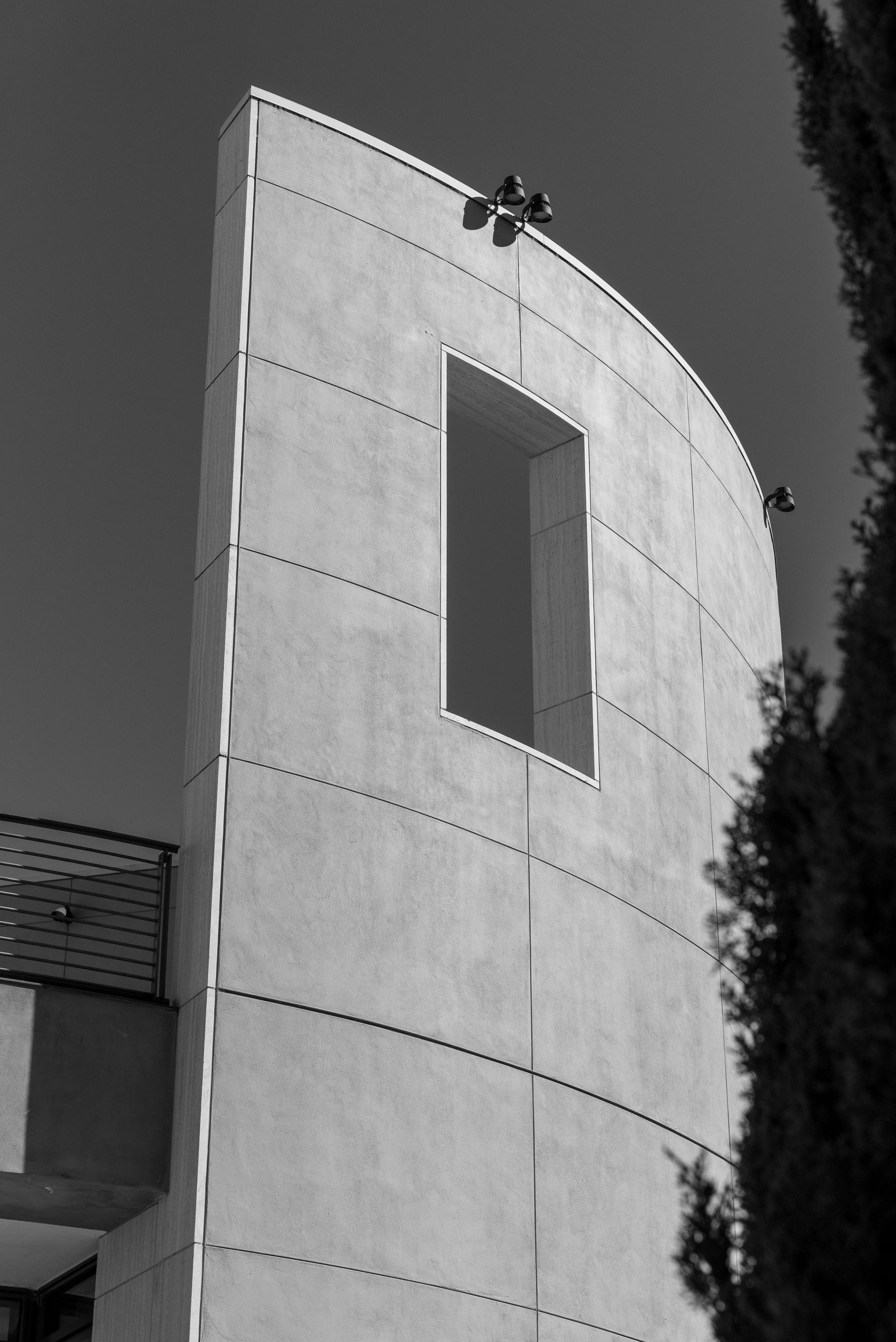 Black and white photo of a smooth, two-story wall which curves away to right. The upper portion of the wall features a single rectangular opening.